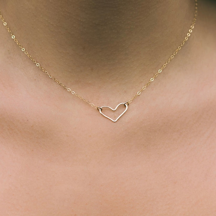 A person wearing a gold-colored open heart necklace on a chain, displayed against a close-up of skin.