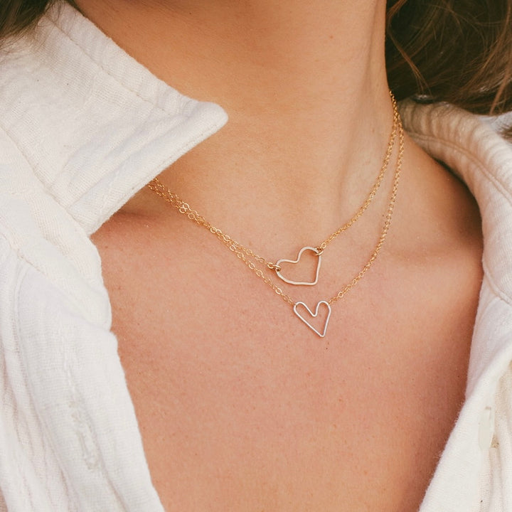 A person wearing a white shirt showcasing a gold-colored necklace with two open heart shapes layered on top of each other.