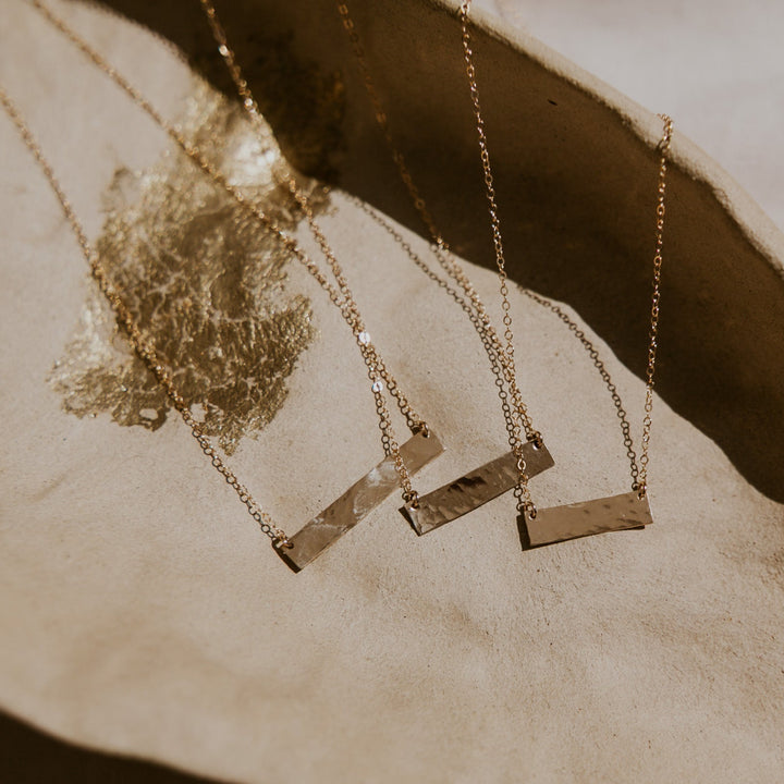 Three gold necklaces with rectangular pendants on a textured beige surface.