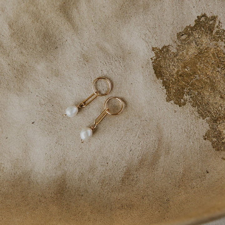 Gold and pearl earrings on a textured beige surface