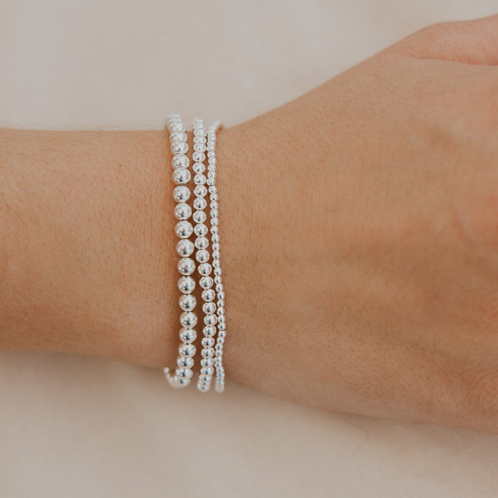 Hand wearing multiple silver beaded bracelets and rings on a neutral background