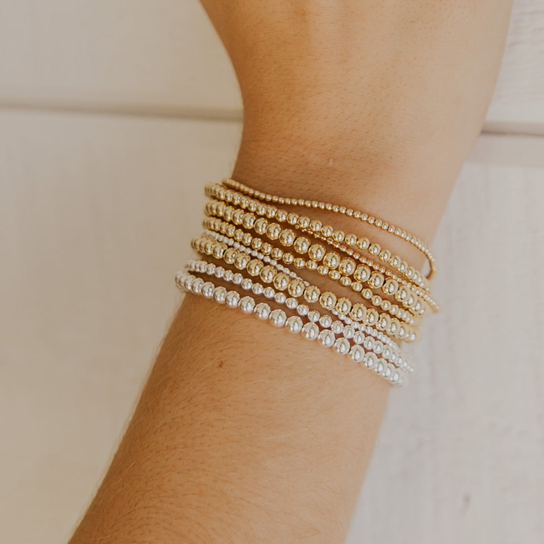 Close-up of a wrist wearing multiple gold and silver beaded bracelets on a neutral background