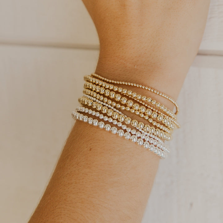 Close-up of a wrist wearing multiple gold and silver beaded bracelets on a neutral background