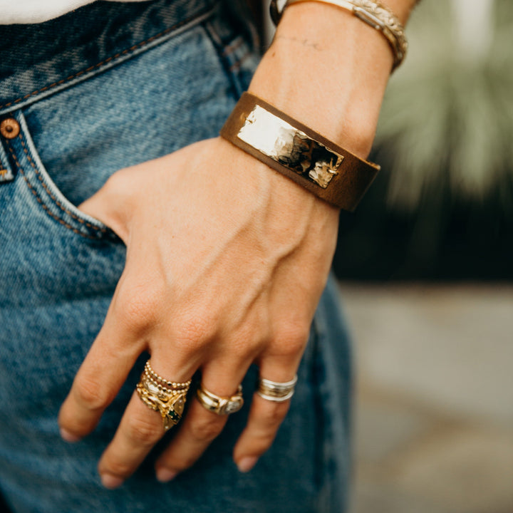 Hand wearing a brown leather bracelet with metallic accents, gold rings, and denim jeans.
