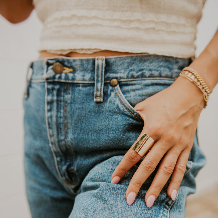 Person wearing blue jeans and a beige top with a gold ring on a neutral background