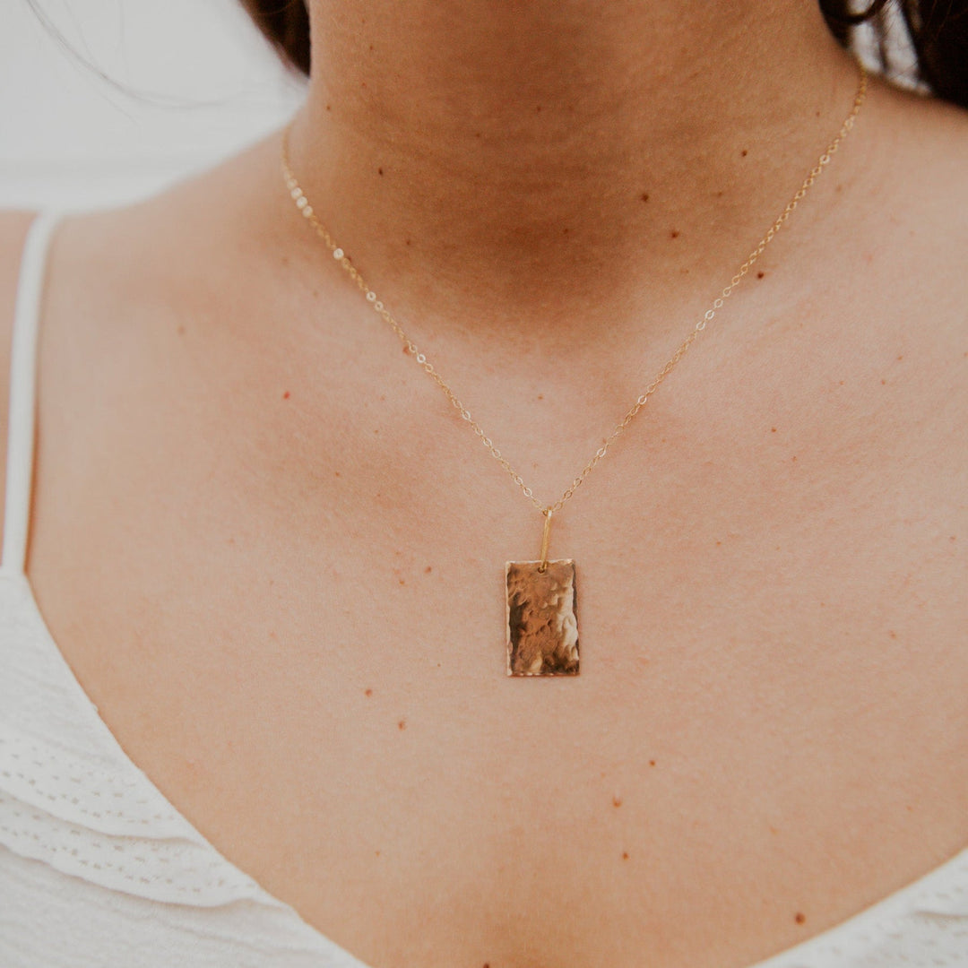 Gold necklace with a rectangular pendant on a person wearing a white top.