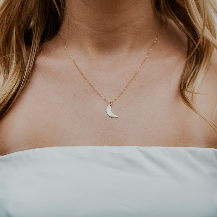 Person wearing a gold necklace with a crescent moon pendant on a blurred background