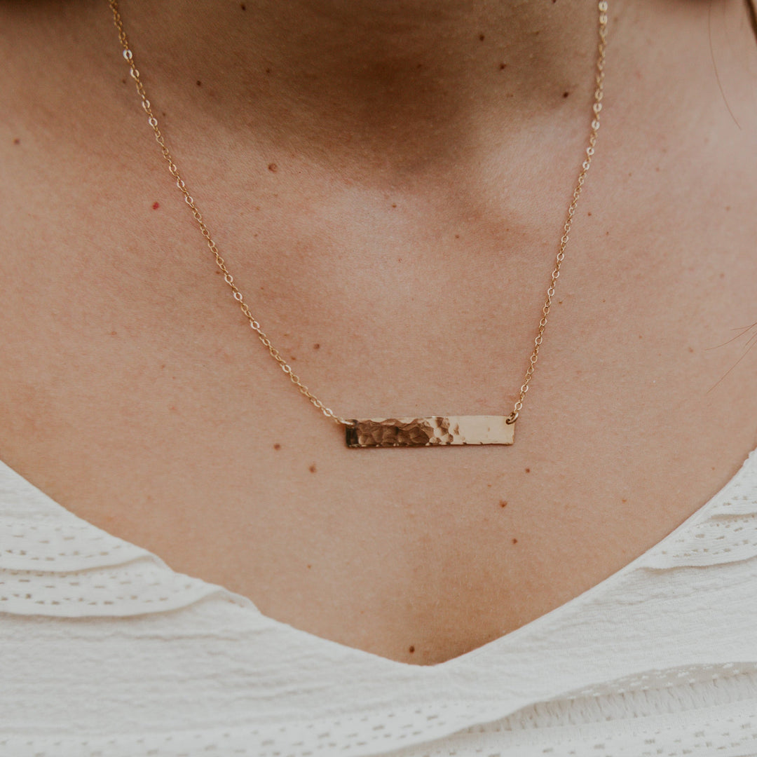Gold necklace with a bar pendant on a person wearing a white top.