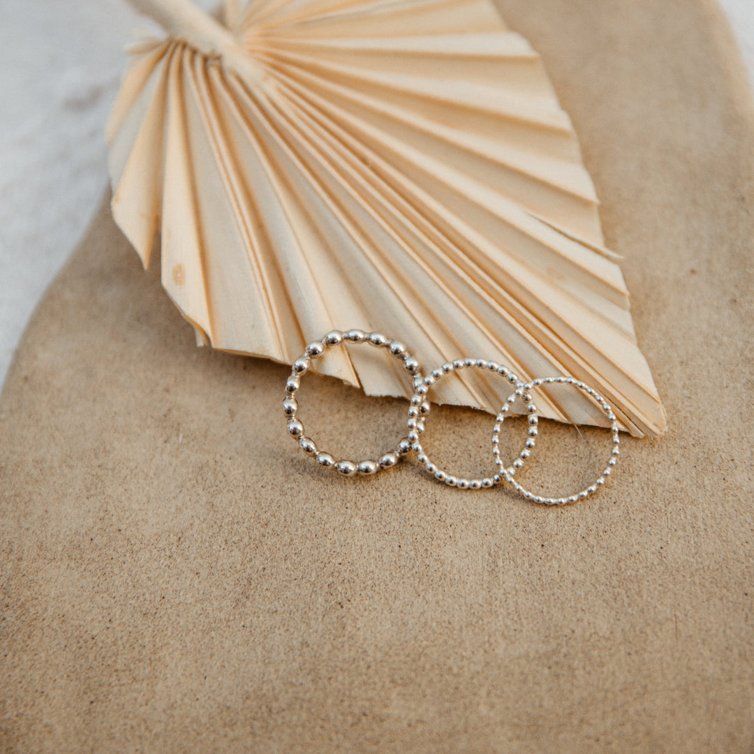 Three beaded silver rings on a textured surface with a fan-shaped object in the background.