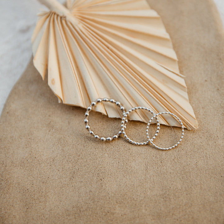 Three beaded silver rings on a textured surface with a fan-shaped object in the background.