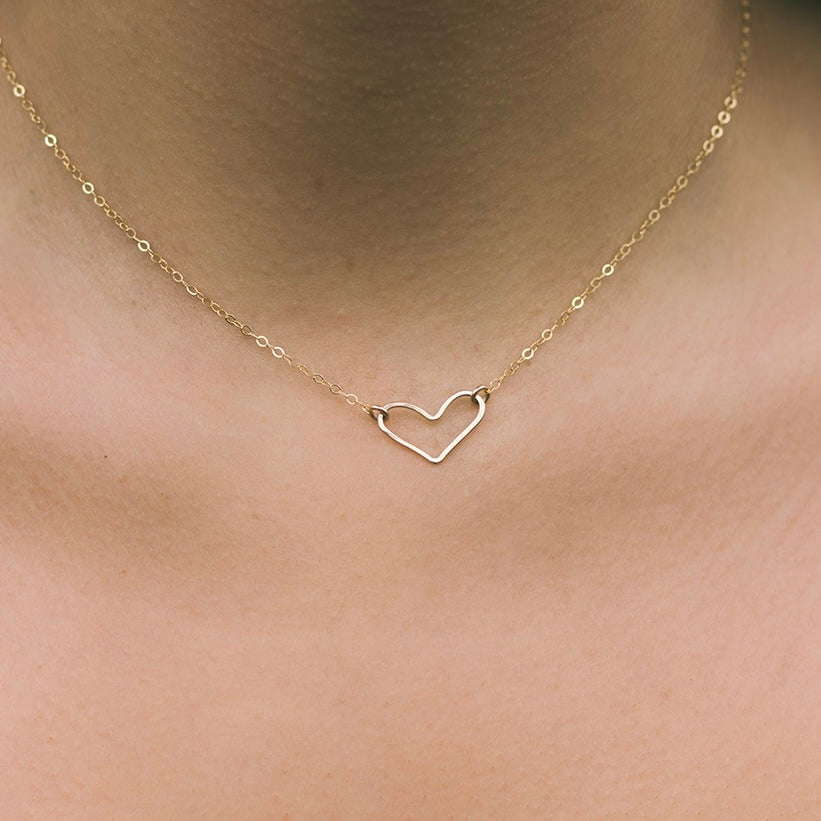 A person wearing a gold-colored open heart necklace on a chain, displayed against a close-up of skin.