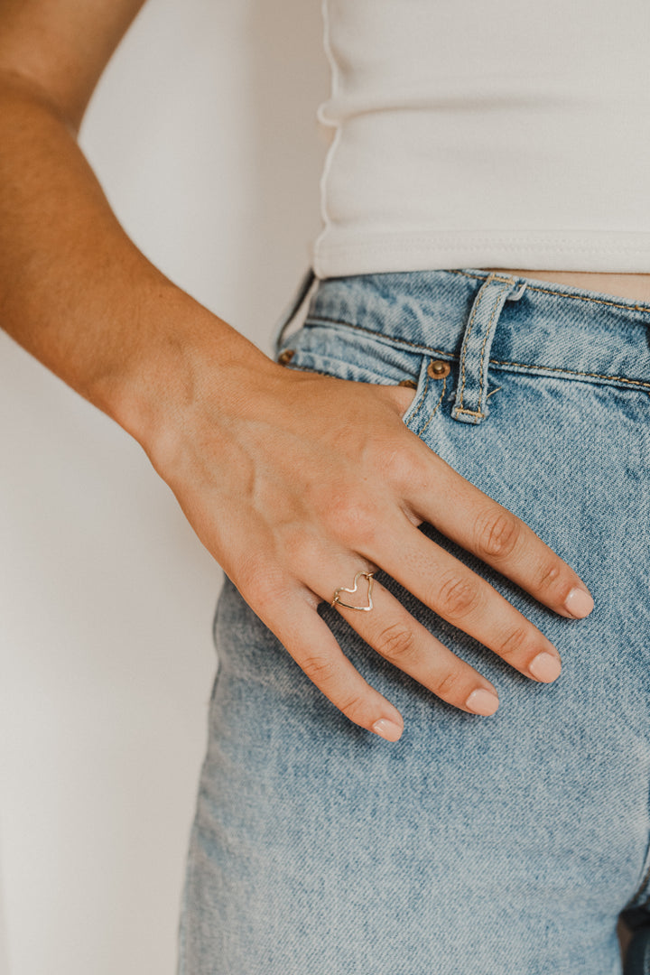 Hand wearing a gold heart shaped ring on a plain background