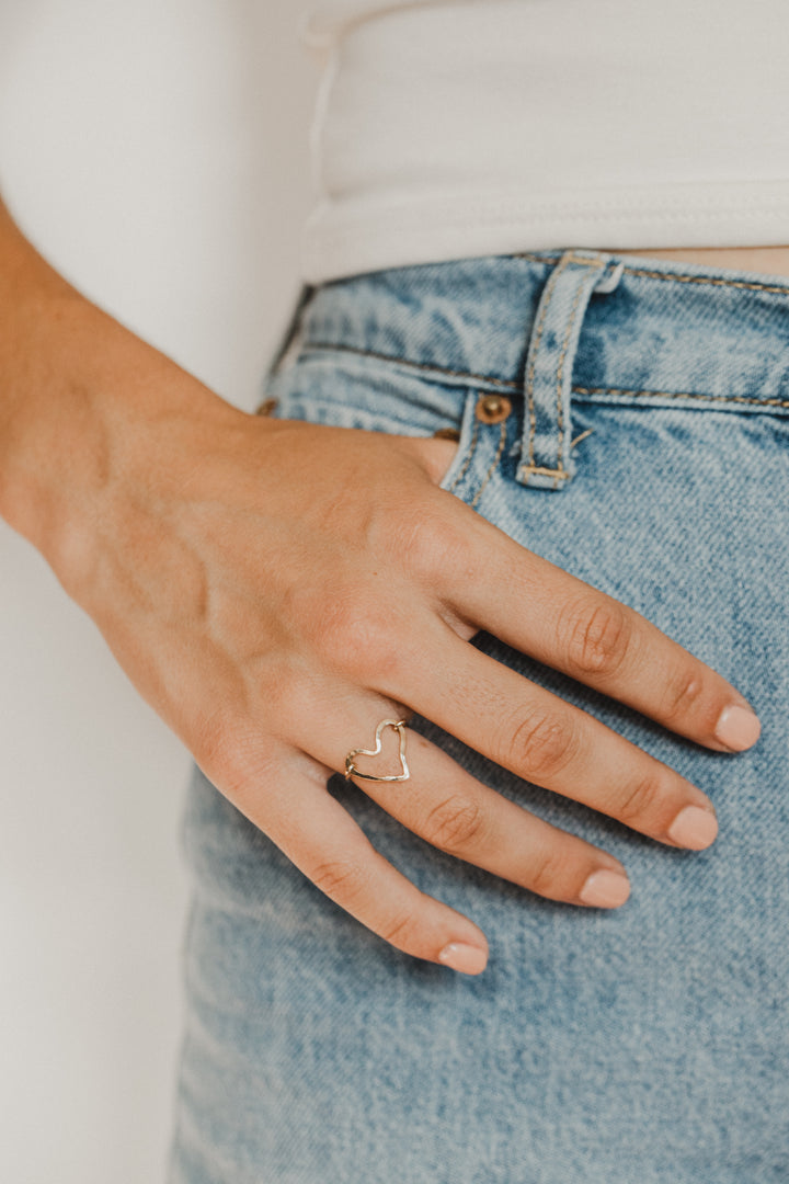 Hand wearing a gold heart-shaped ring on a blurred background