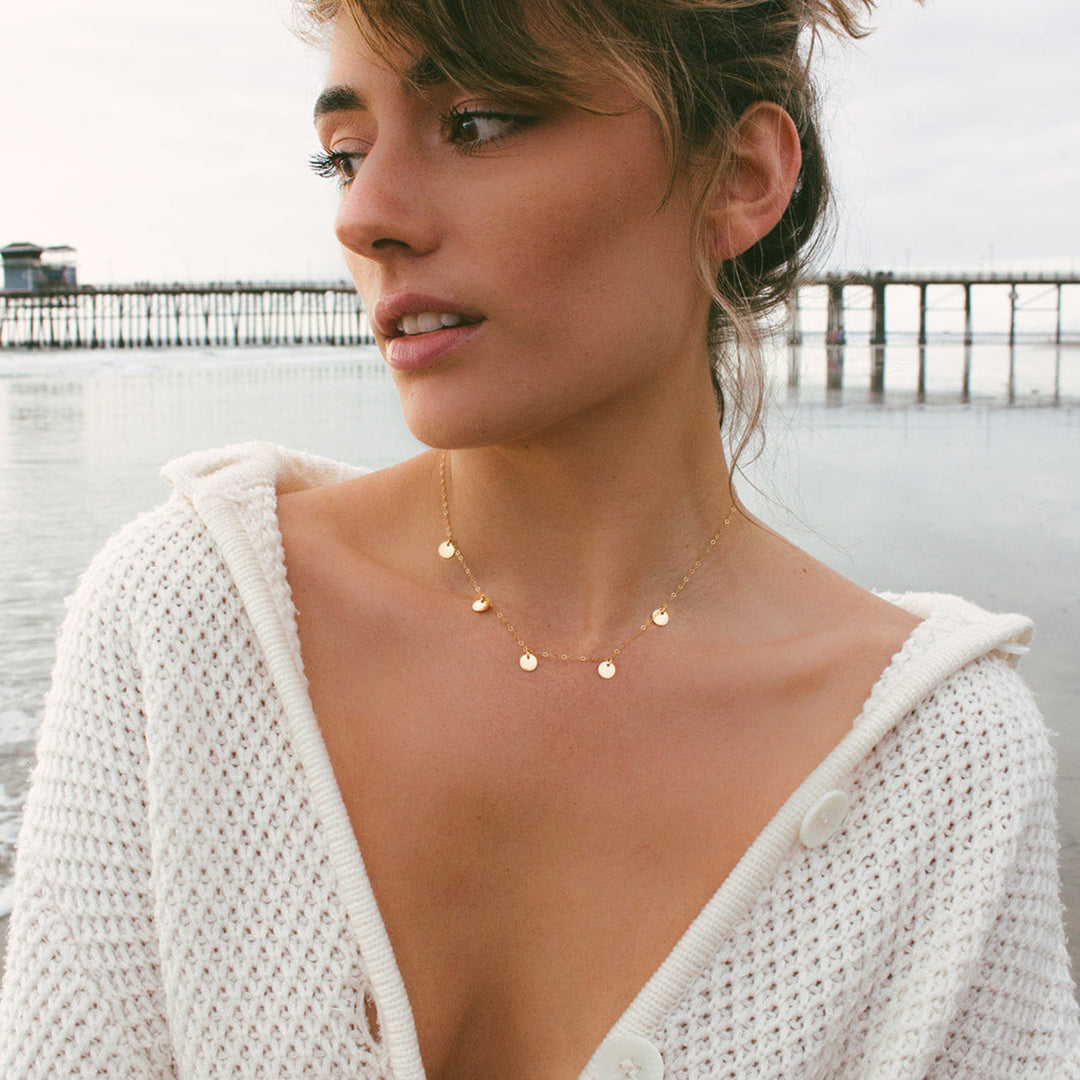 Woman wearing a white knit cardigan and gold disc necklace by a body of water with a pier in the background