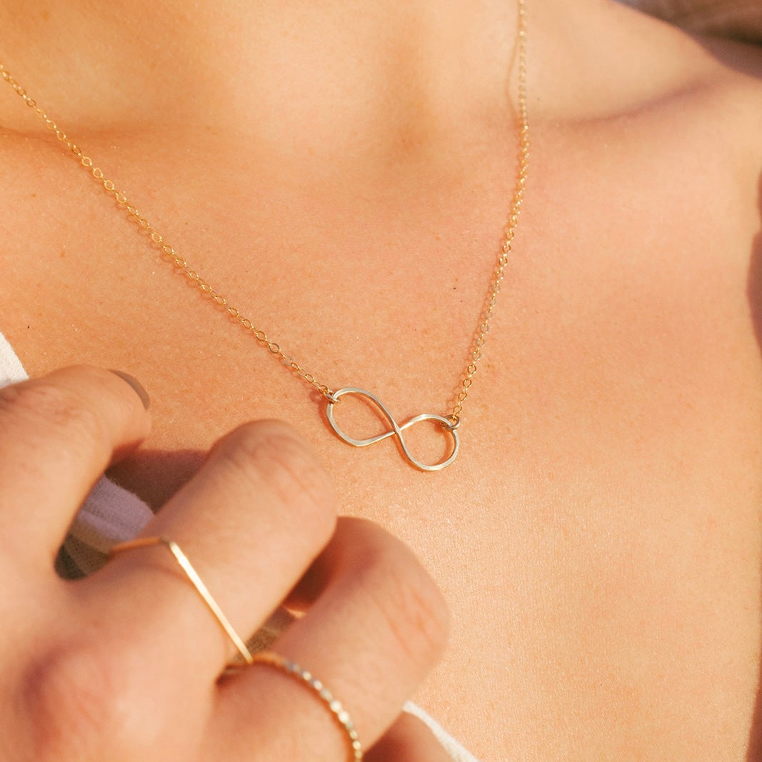 A close-up image of a woman's neck wearing a gold infinity necklace, with a focus on the necklace's texture and shine.