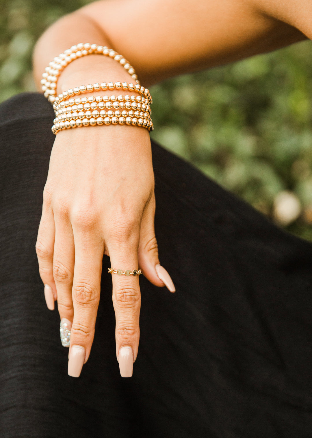 Hand wearing gold bracelets and gold chain link rings with a blurred natural background