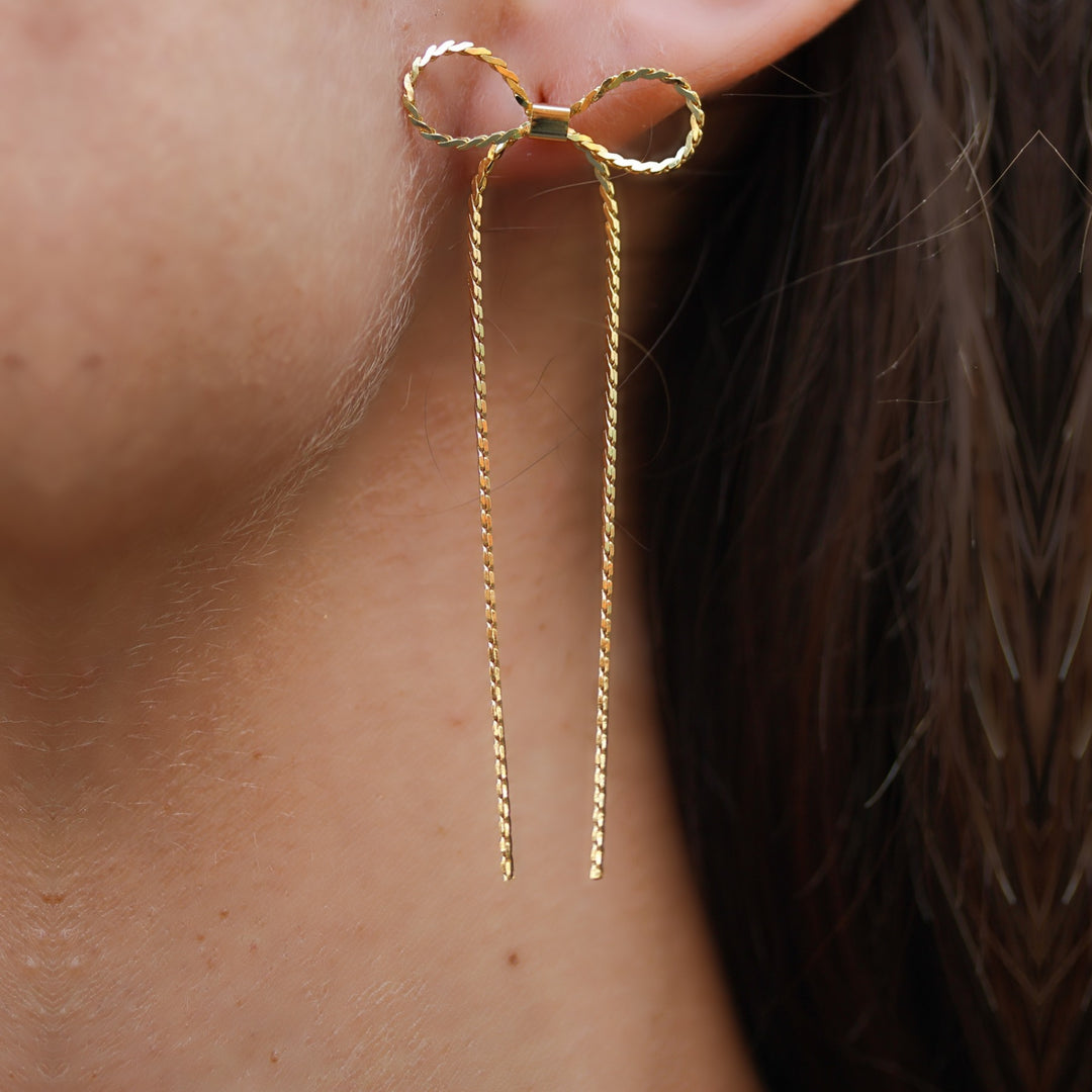 Close-up of the Long Dangle Coco Bow Earring, a twisted gold bow with long ribbon tails, gold plated over brass