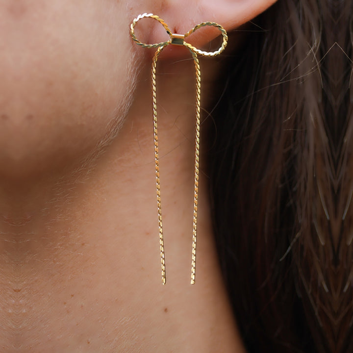 Close-up of the Long Dangle Coco Bow Earring, a twisted gold bow with long ribbon tails, gold plated over brass