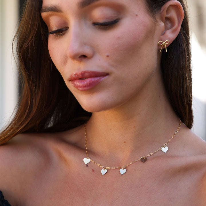 Model looking down wearing the Short Coco Bow Earring with a gold heart charm necklace