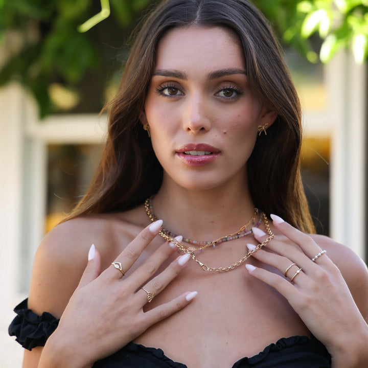 Model wearing Short Coco Bow Earrings with layered tourmaline and gold chain necklaces and stacked rings
