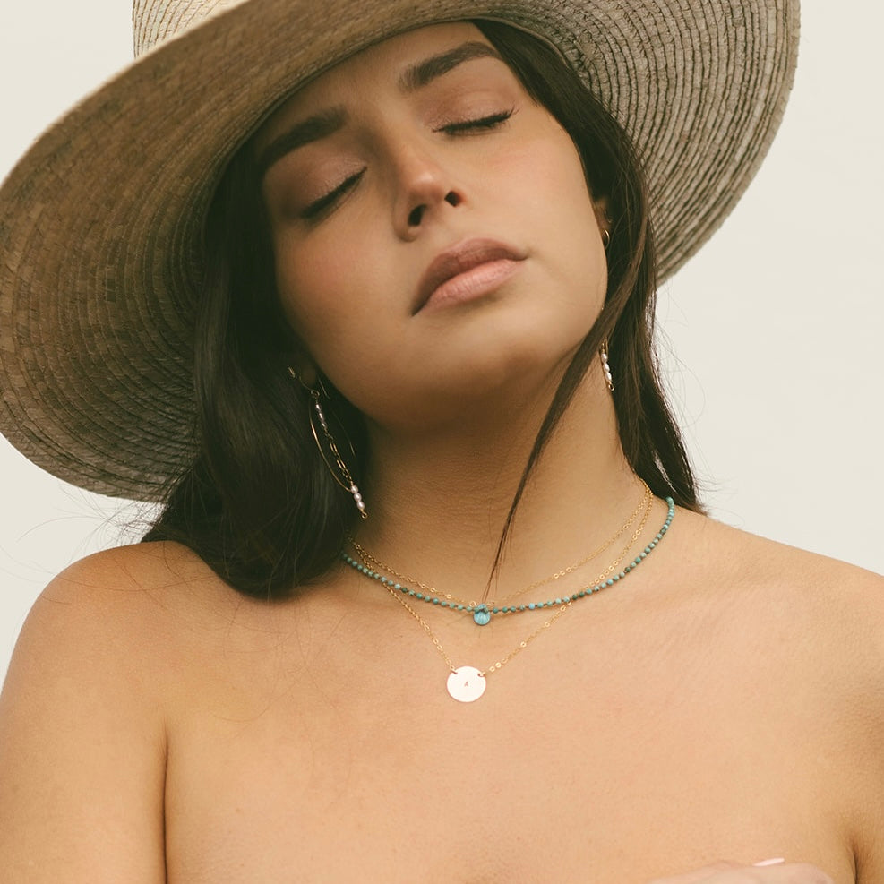 Woman wearing a Teressa Lane Jewelry gold disc necklace for women with layered turquoise beads and a wide-brimmed hat