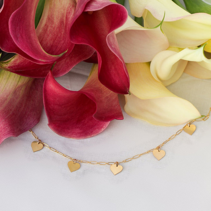 Heart necklace with five small gold filled heart dangles on fine paperclip chain, laid in front of pink and cream calla lilies