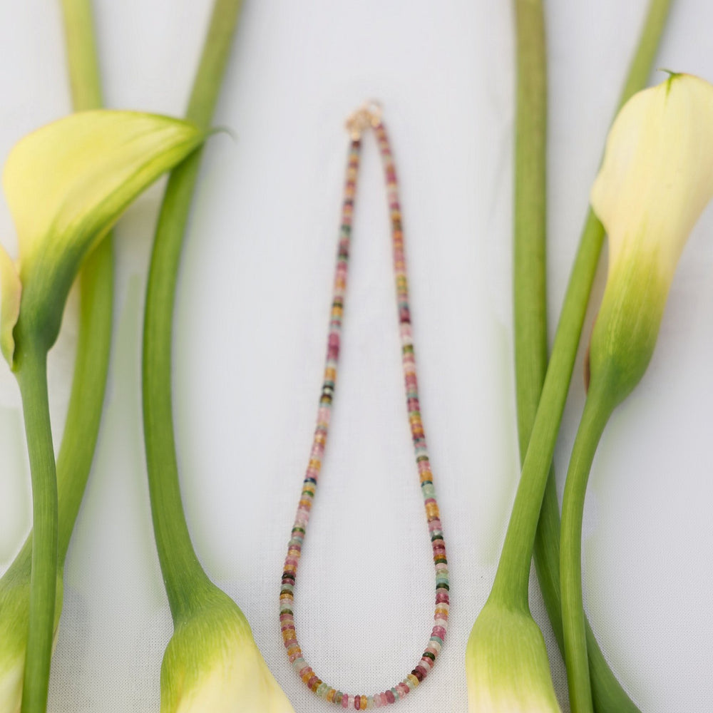 Joy necklace, a strand of faceted multicolor tourmaline heishi beads with a gold filled clasp, laid between cream calla lilies