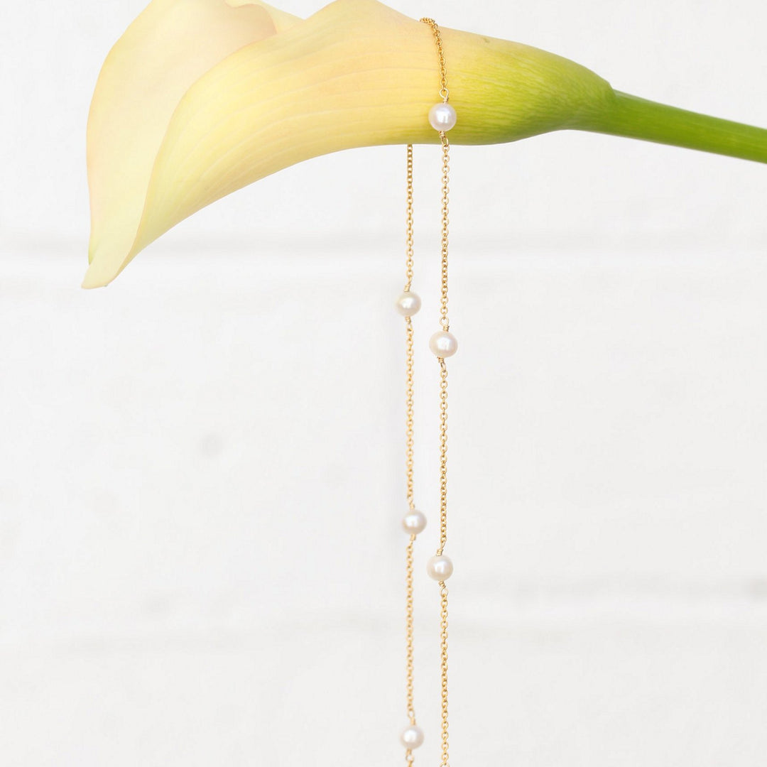 Stella gold filled pearl station chain draped over a cream calla lily against a white brick wall